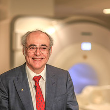 a man with grey hair wearing a grey suit , white shirt and red tie, stands before the blurred out image of an mri machine