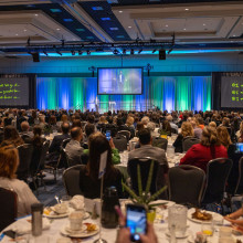 A full ball room of event attendees watching the keynote speaker on stage