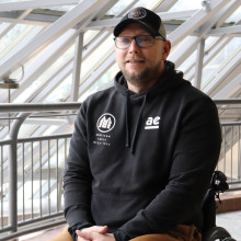 Chris smiles for the camera in a black hoodie, hat and brown pants